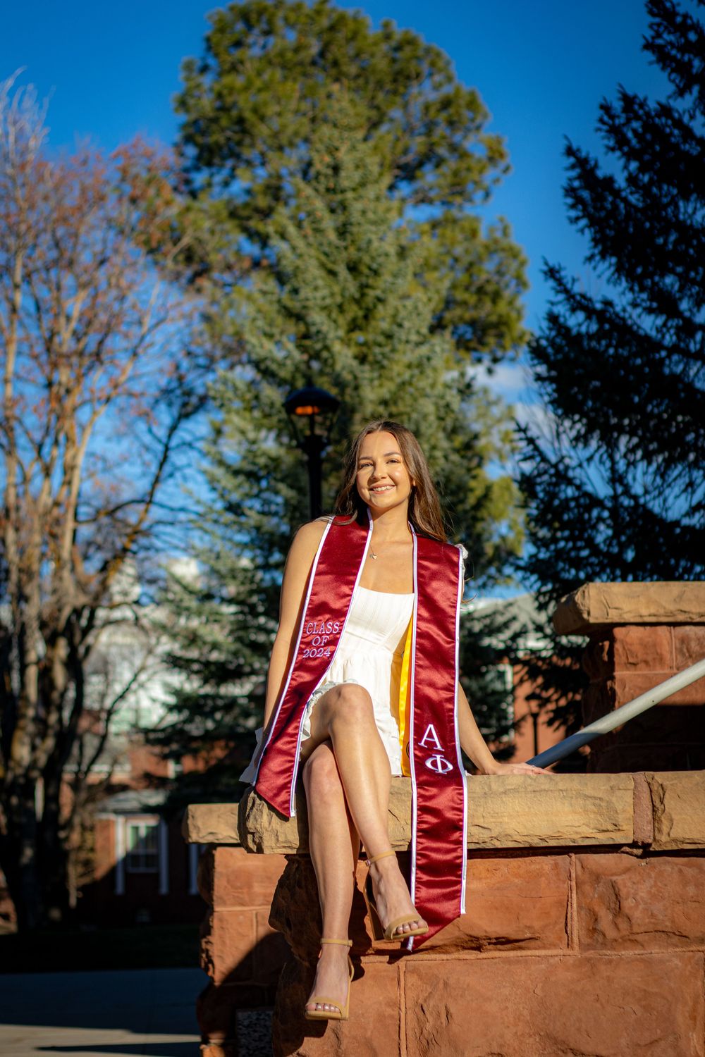 Graduate in red stole poses on stone steps with tall pine tree in background.