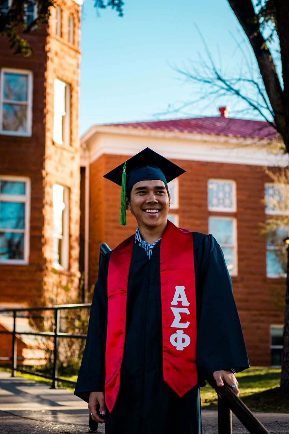 Graduate wearing black cap and gown with red fraternity stole poses in front of brick campus building.