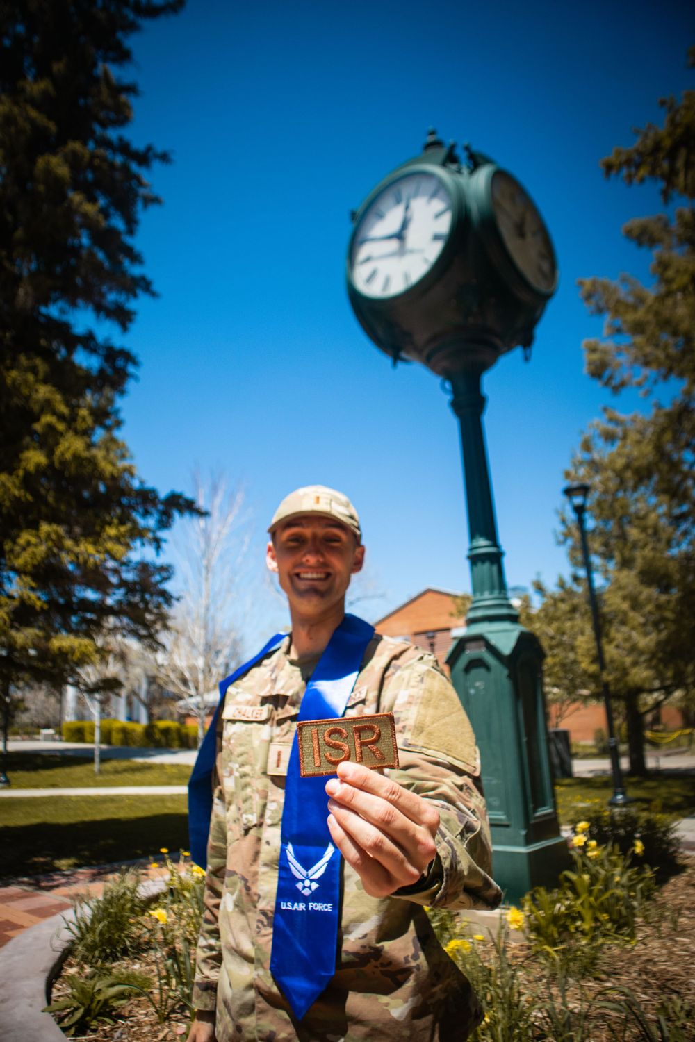 Military servicemember in tan jacket holds up graduation stole near vintage campus clock tower.