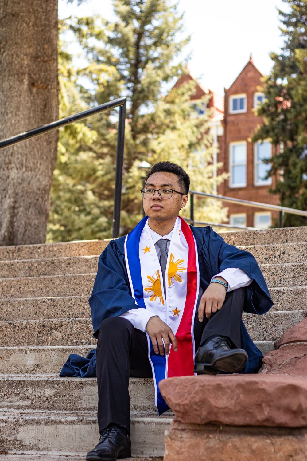 Graduate in blue cap and gown with decorated stole sits on stone steps near historic building.