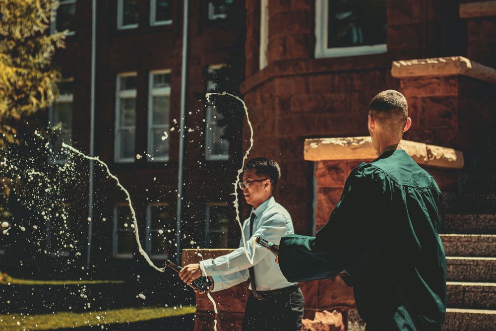 Students celebrating graduation with water spray on campus steps near brick building.