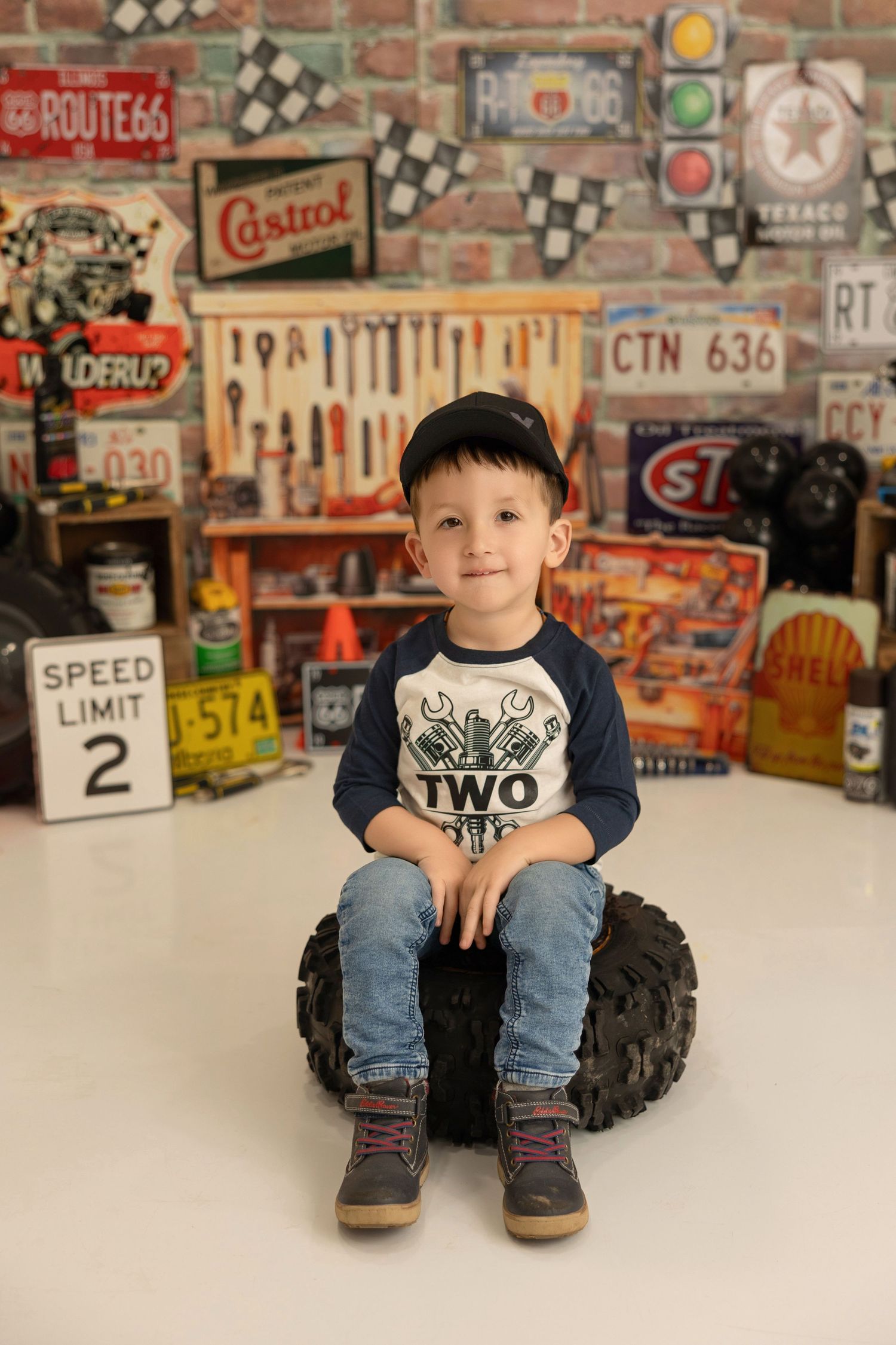A young child sits on a large tire in front of a retro garage-themed backdrop with vintage signs and tools.