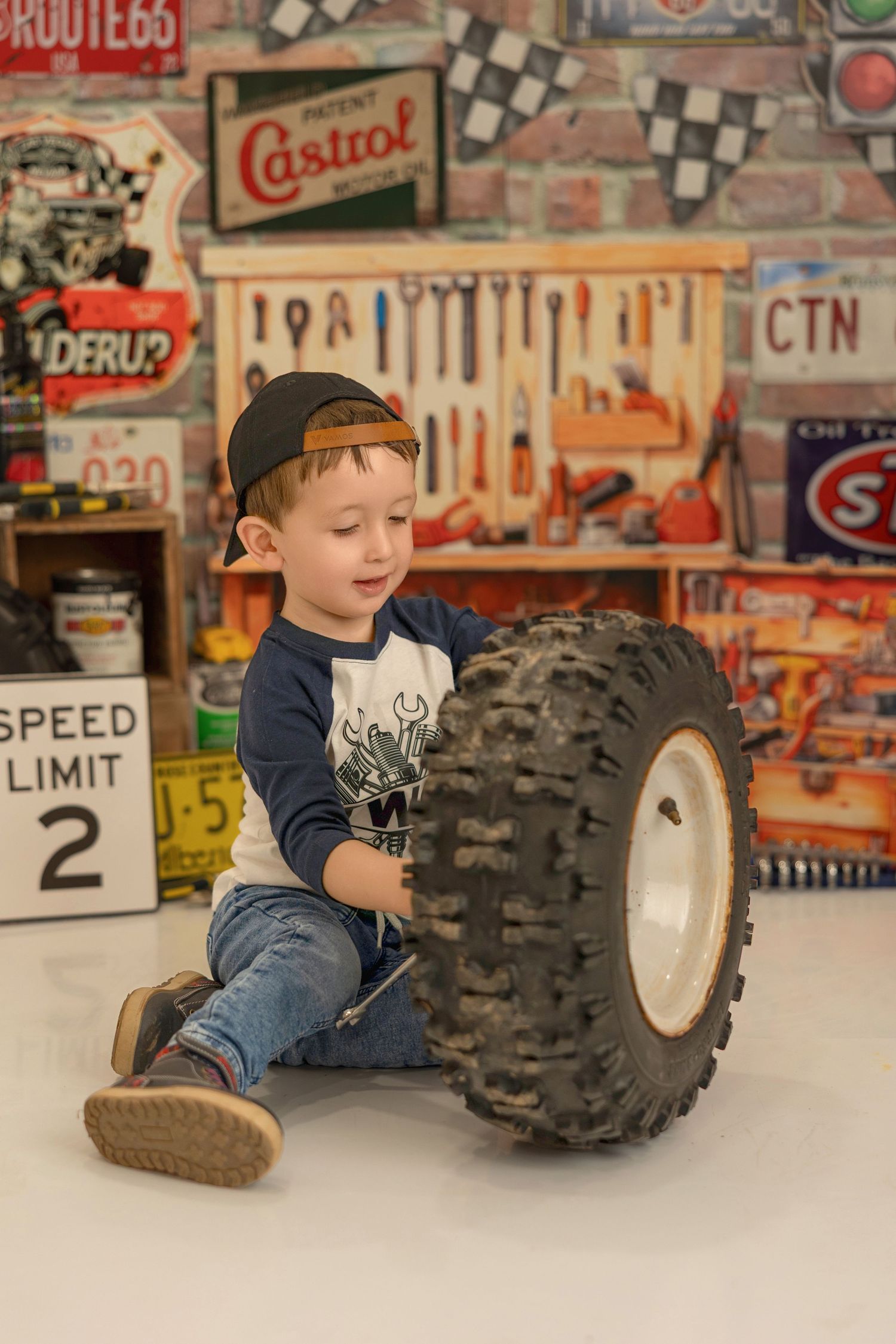 Young child playing near a large ATV tire in a room decorated with vintage automotive signs and memorabilia.