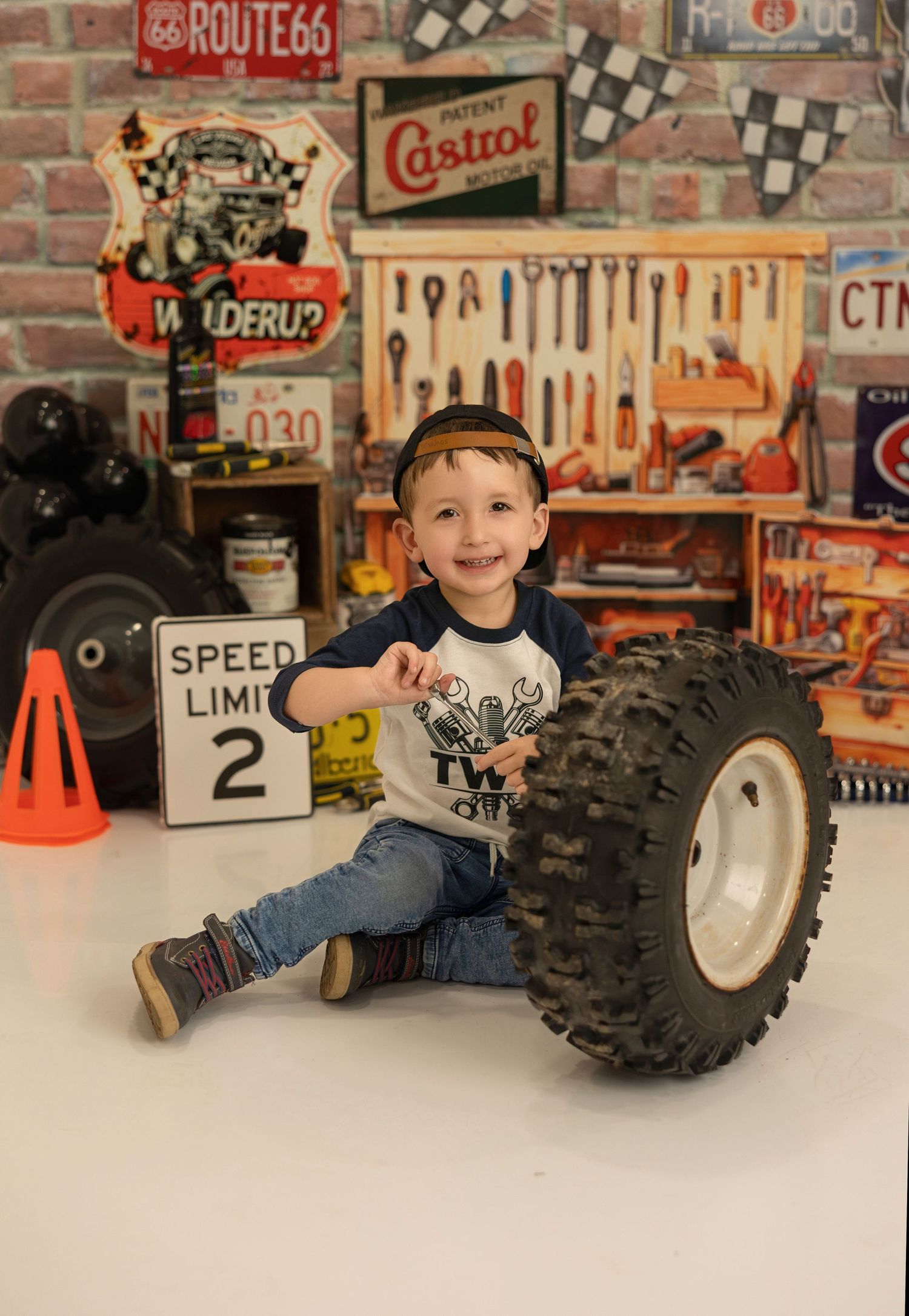 Child smiling playfully beside large off-road vehicle tire in a retro-styled garage setting.
