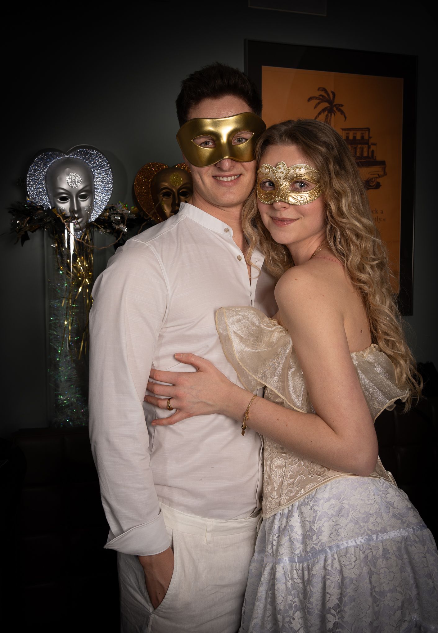 A couple in elegant white attire wearing gold masquerade masks pose together at a formal event.