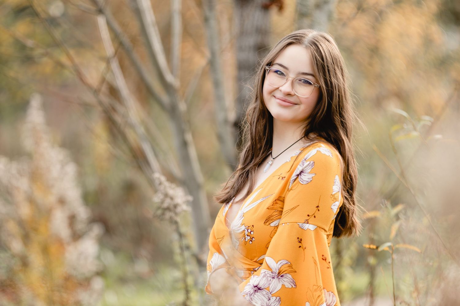 A young woman wearing a yellow floral dress poses outdoors during autumn with a gentle smile.