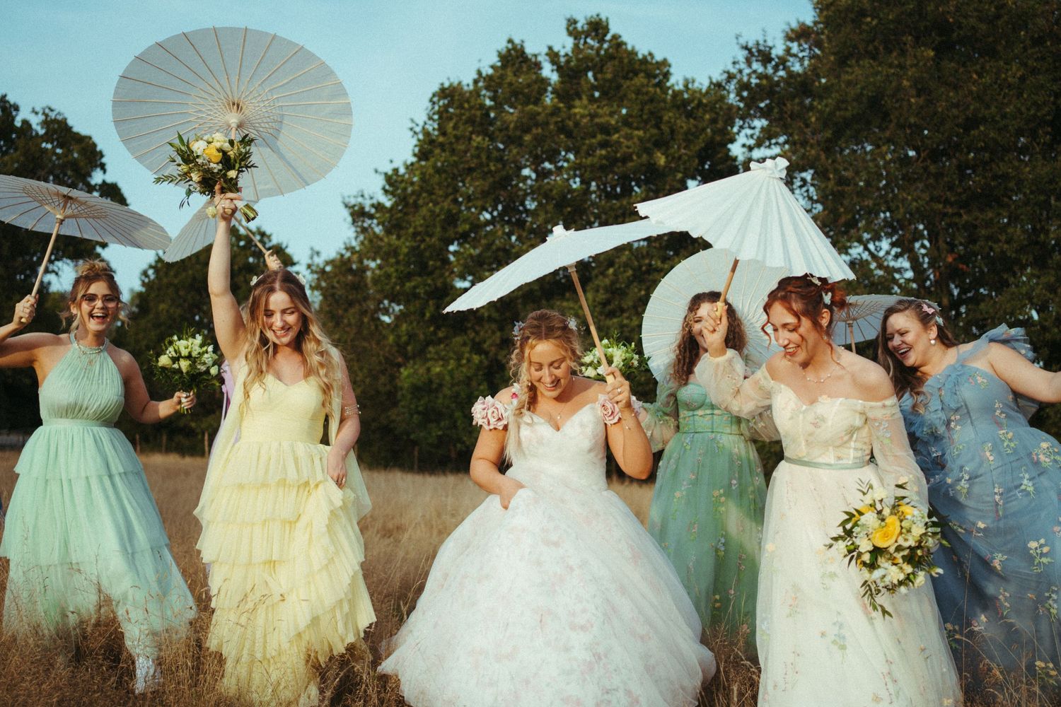 A group in whimsical pastel dresses poses with decorative parasols in a sunlit outdoor field.
