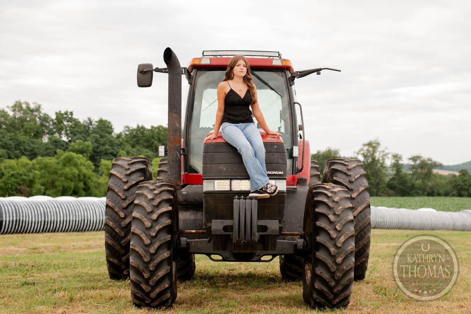 Person in black top and jeans poses confidently on large red farm tractor.
