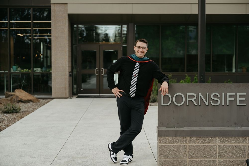Graduate in black cap and gown poses casually leaning against building exterior with Dornsife signage.