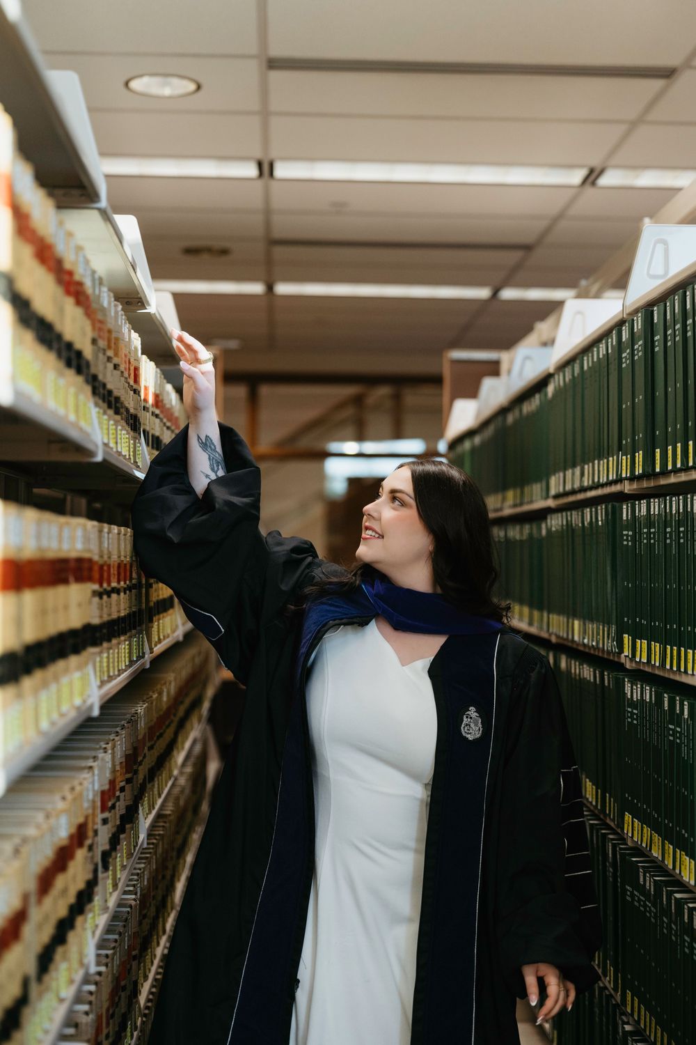 A woman in graduation regalia browses shelves of green law books in a library aisle.