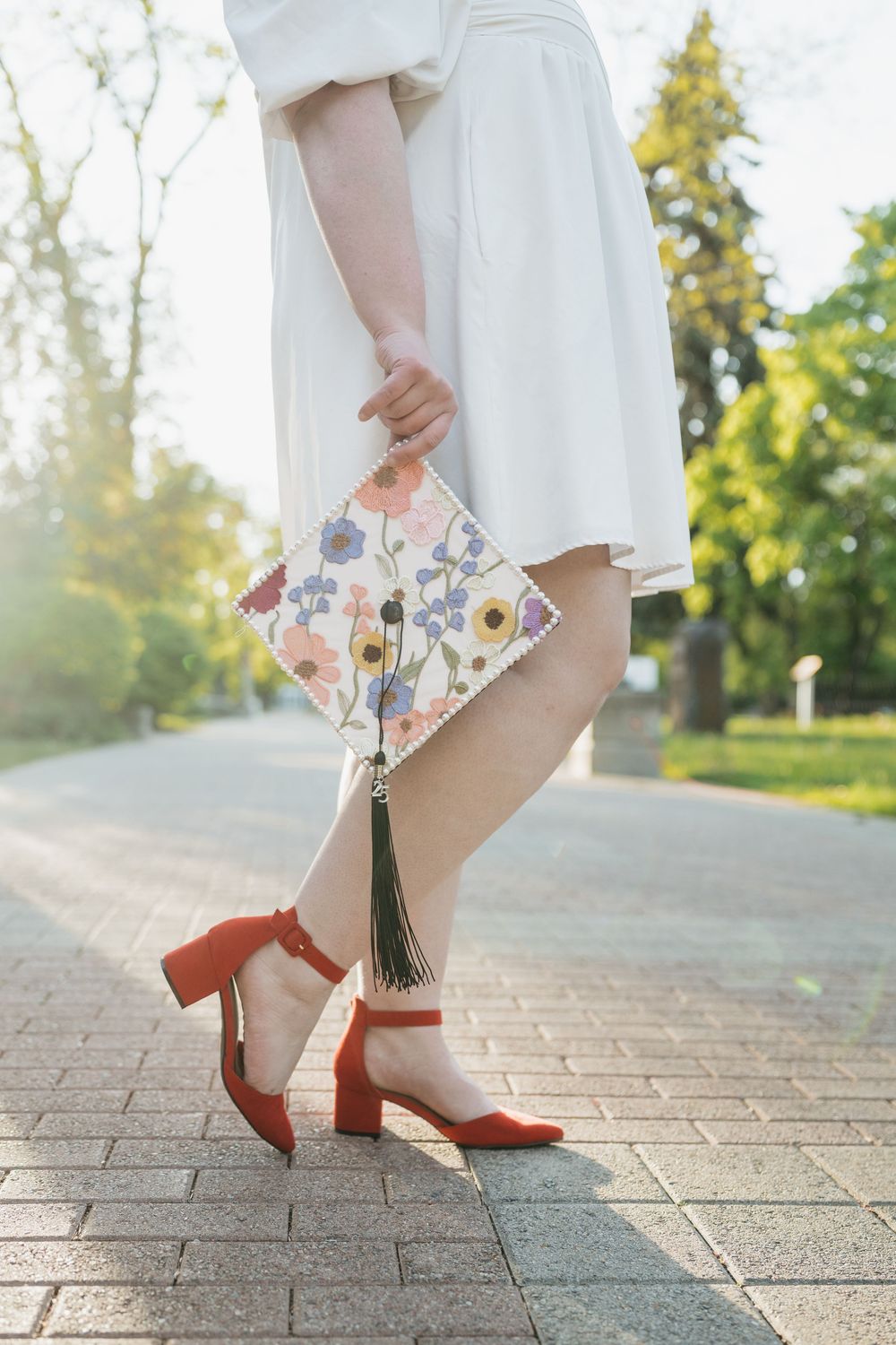 Woman wearing white dress and red block heel shoes holds floral printed clutch purse on brick pathway in sunlit garden.