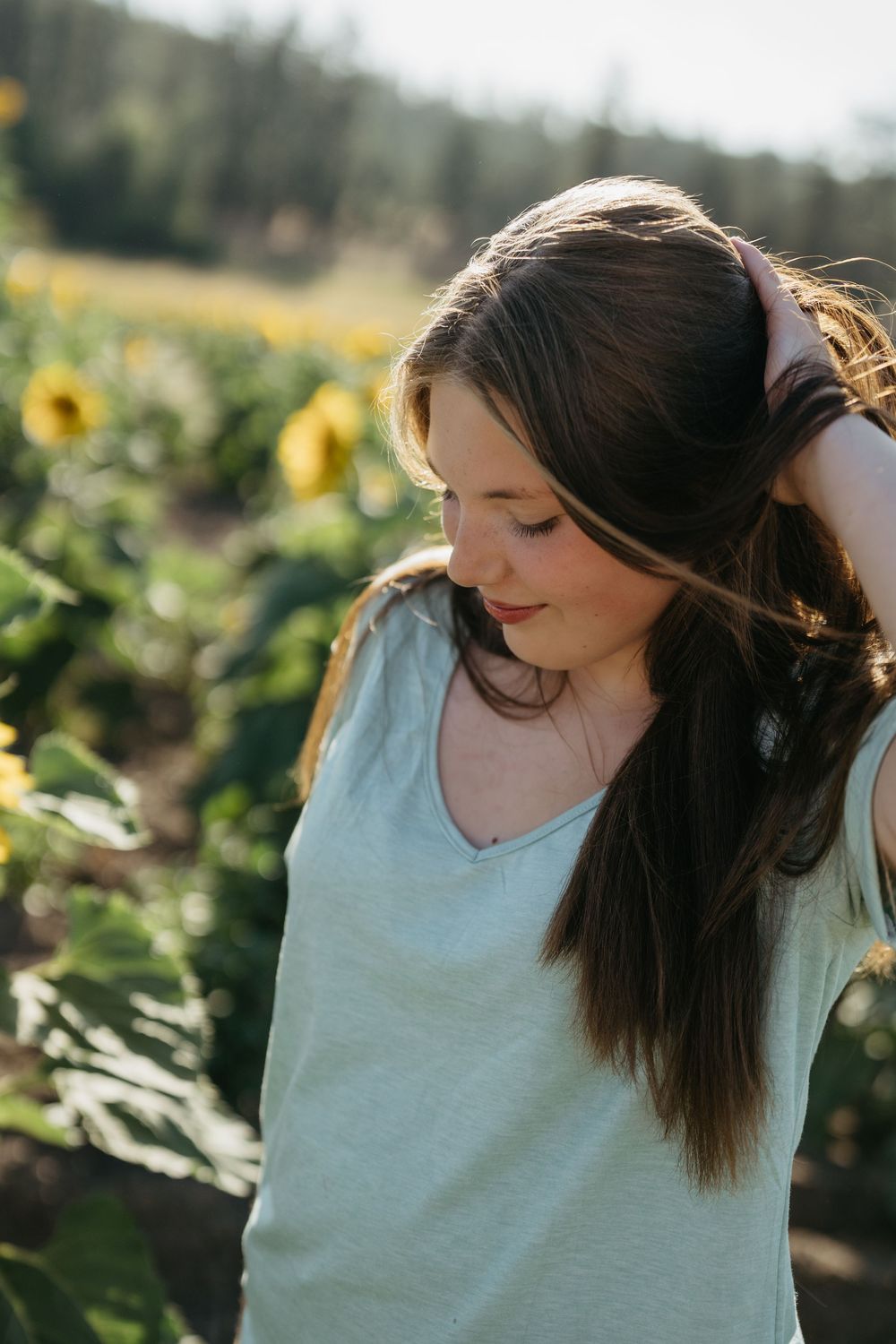 A young woman in a light blue top stands in a sunflower field with sunlight creating a warm summer glow.