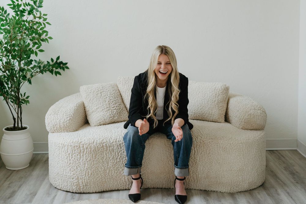 Smiling blonde woman in black blazer and jeans sitting on a cream sherpa sofa in a bright minimalist room.