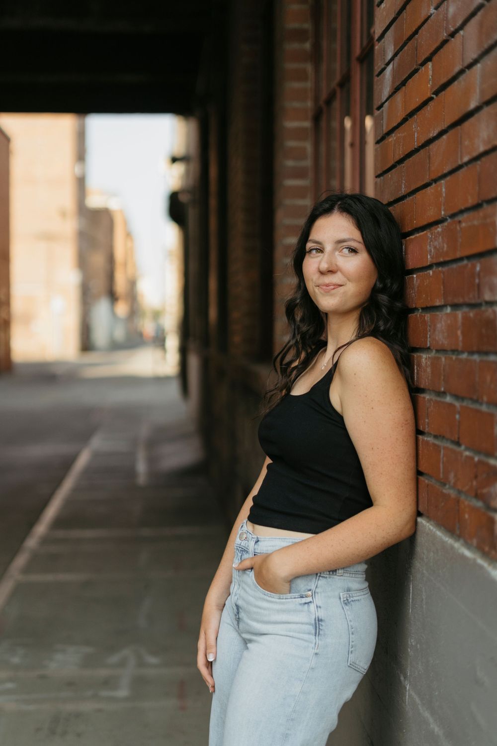 A casual portrait against a brick wall shows a relaxed pose in a black tank top and light jeans in an urban alley.