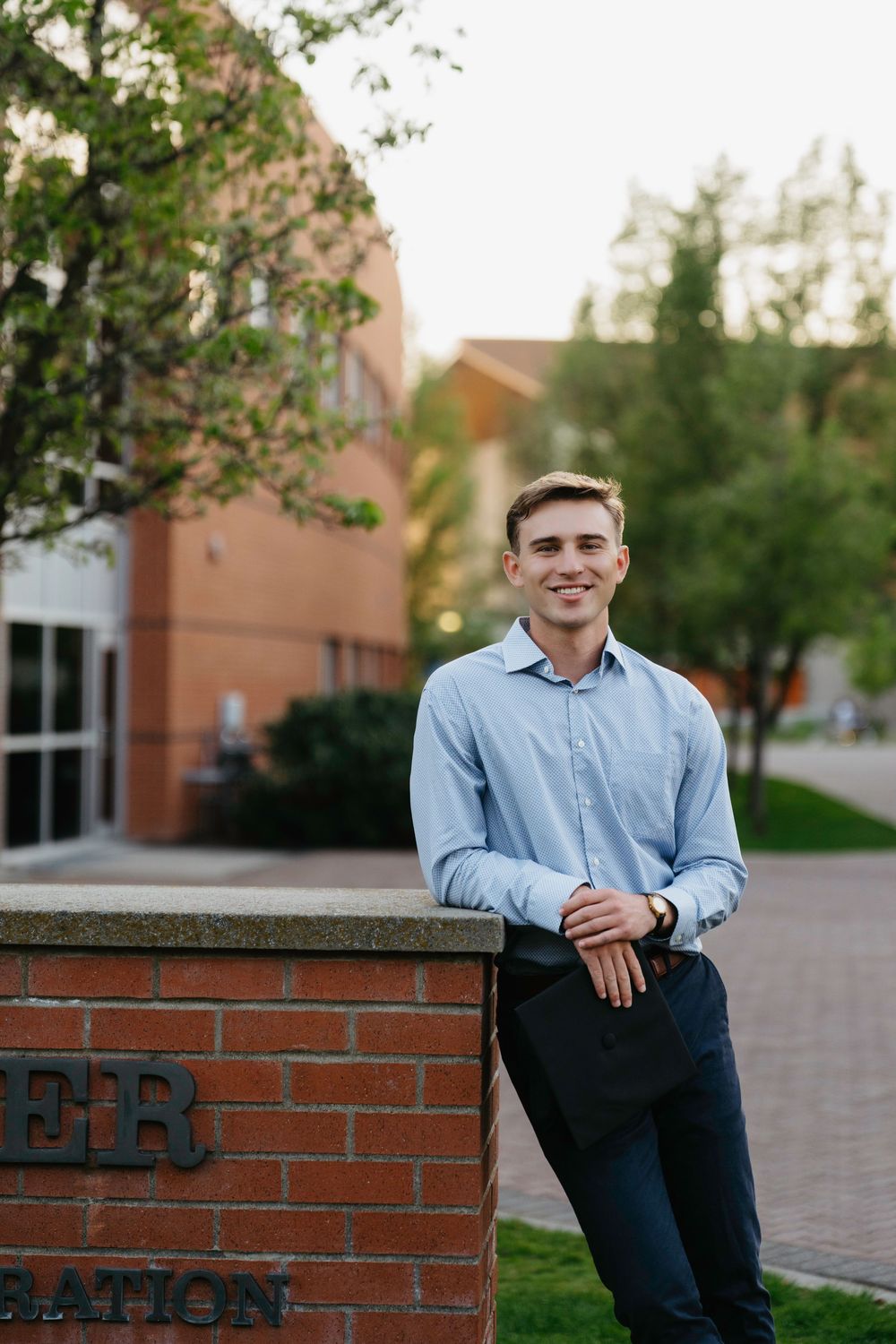 Young man in blue shirt smiling while leaning against a brick sign on a college campus.