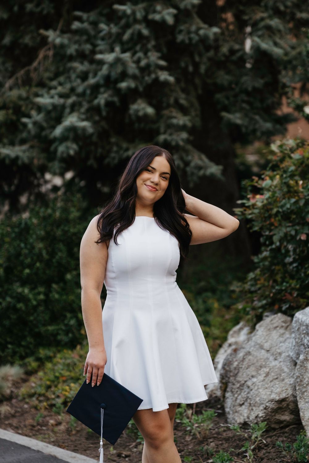 Young woman in white dress holding graduation cap, smiling outdoors near trees and rocks.