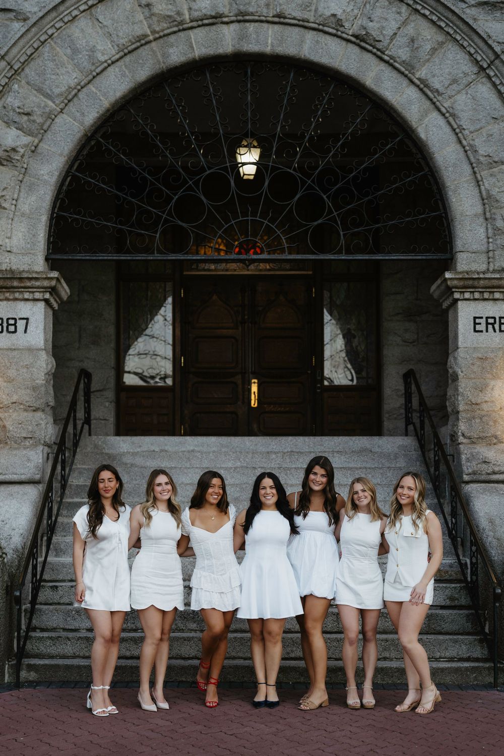 Seven women in white dresses pose together on stone steps in front of an arched building entrance.