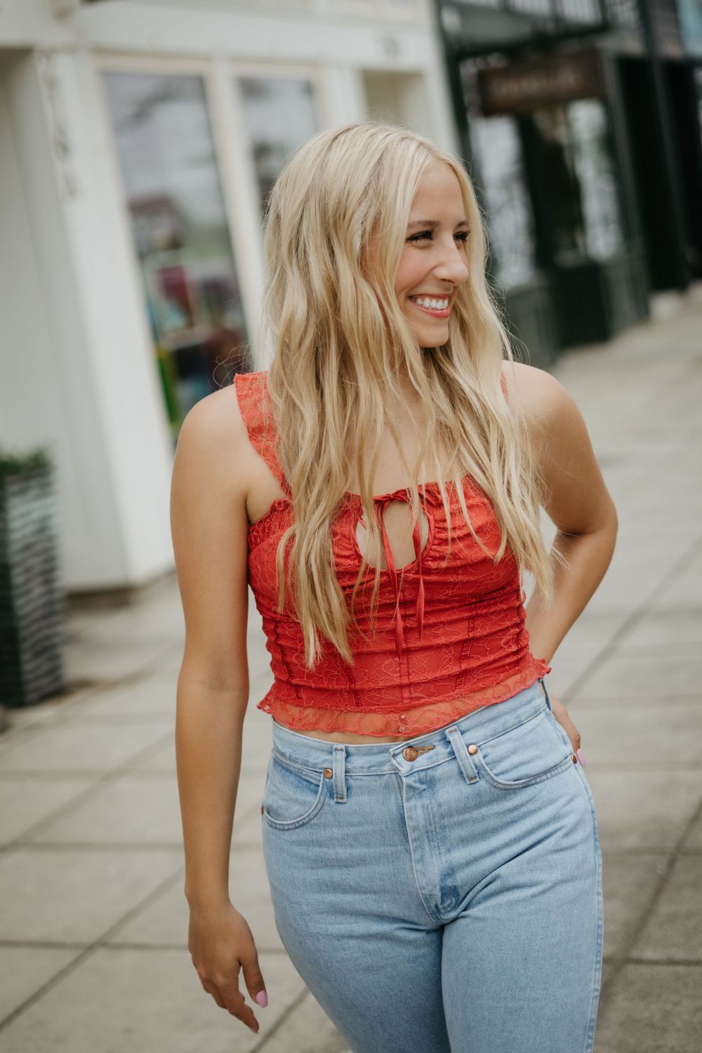 A woman in a red sleeveless top and high-waisted jeans laughs while standing on a city sidewalk.