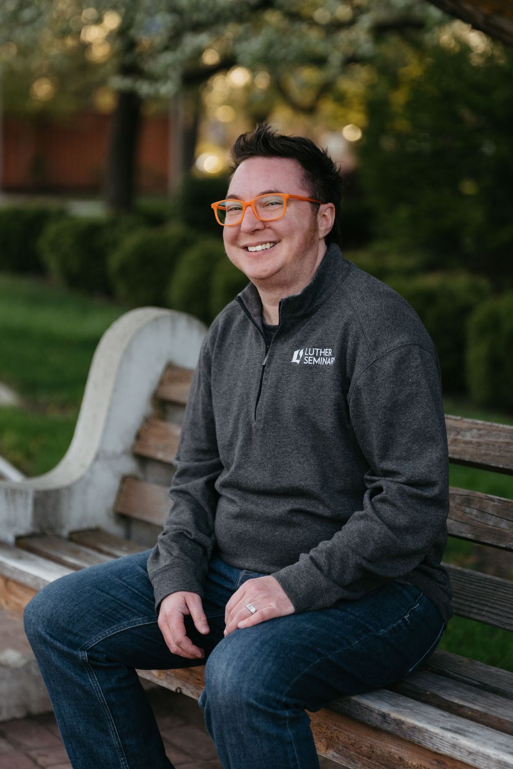 Person wearing orange glasses and grey fleece sitting on wooden park bench smiling outdoors on sunny day.