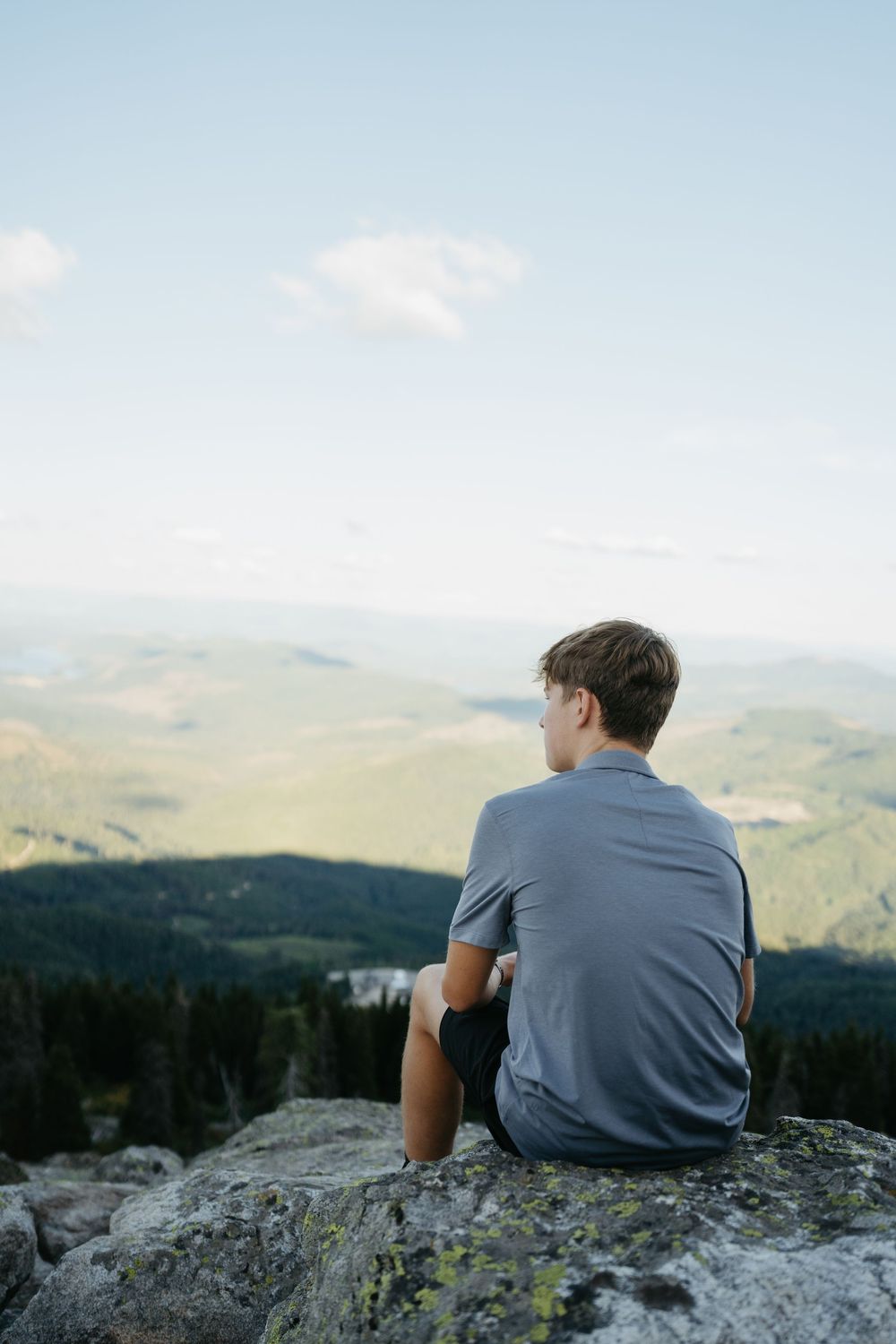 A person in a gray shirt sits on a rocky mountain overlook gazing at the scenic valley and rolling hills below.