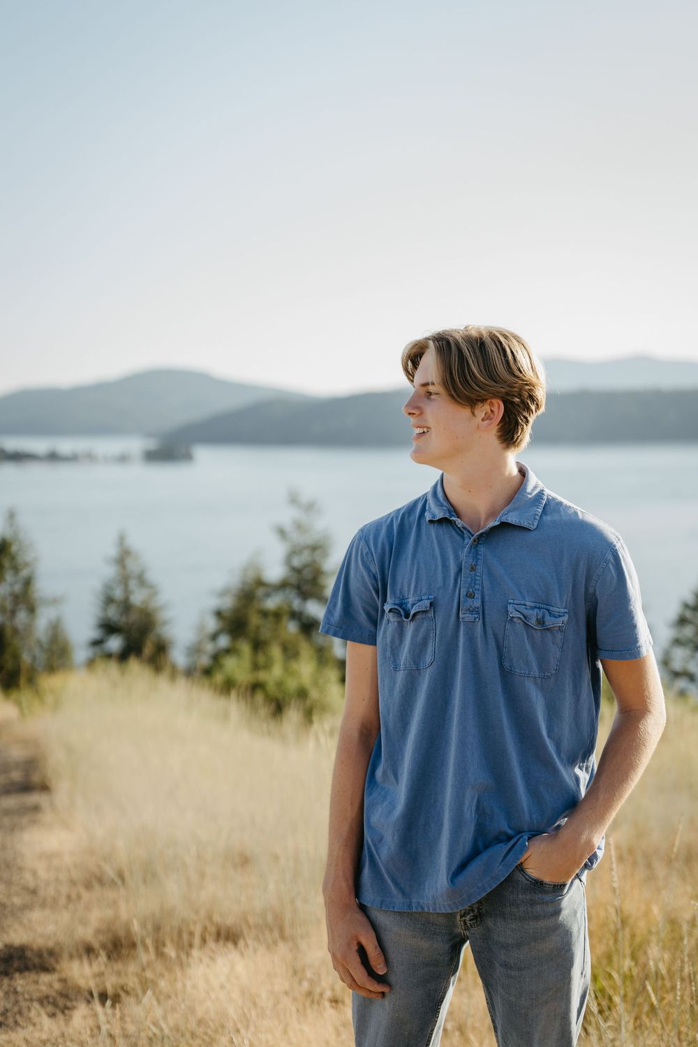 Person in denim shirt stands in golden grass field overlooking scenic mountain lake view on sunny day.