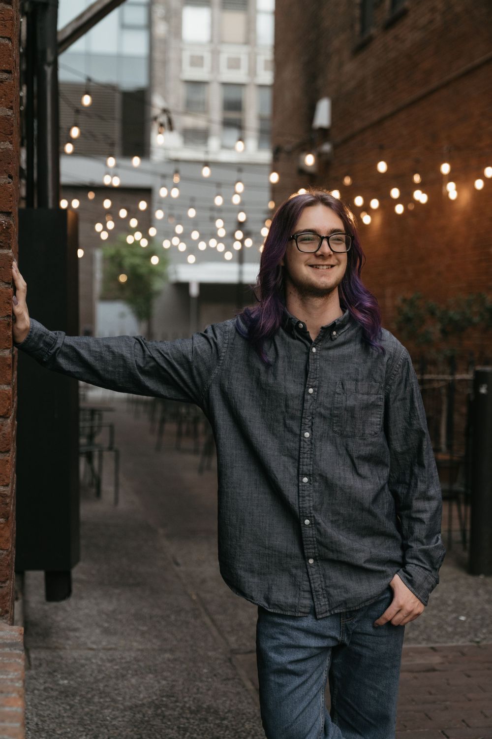 Person in glasses and gray button-up shirt smiling in an alley with string lights overhead in evening light.