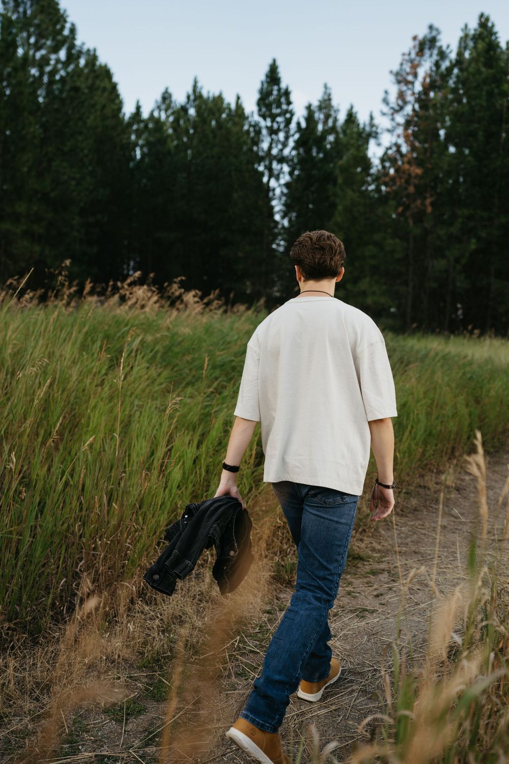 Person in white t-shirt and jeans walking through a tall grass field with pine trees in the background.