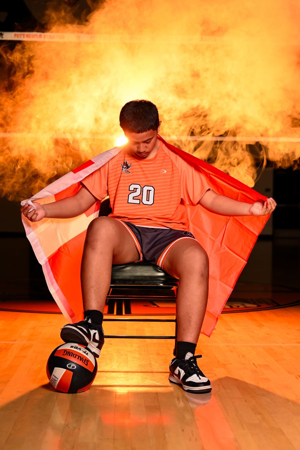 Basketball player stretches in orange jersey on hardwood floor with dramatic lighting.