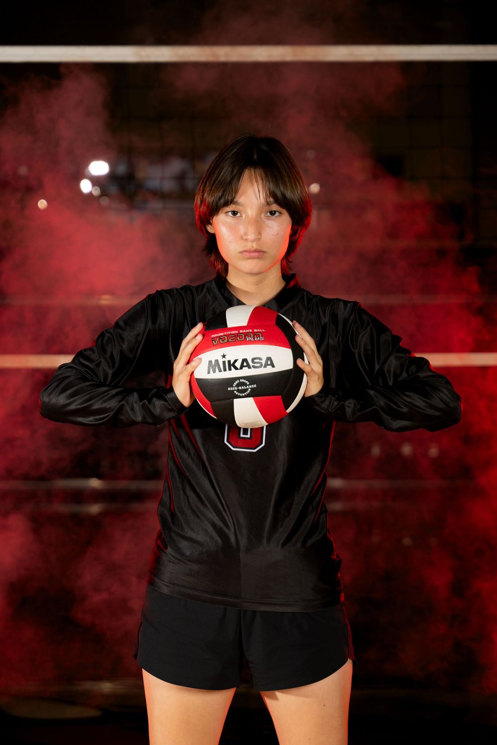 Player in black jacket poses with volleyball against red smoky background.