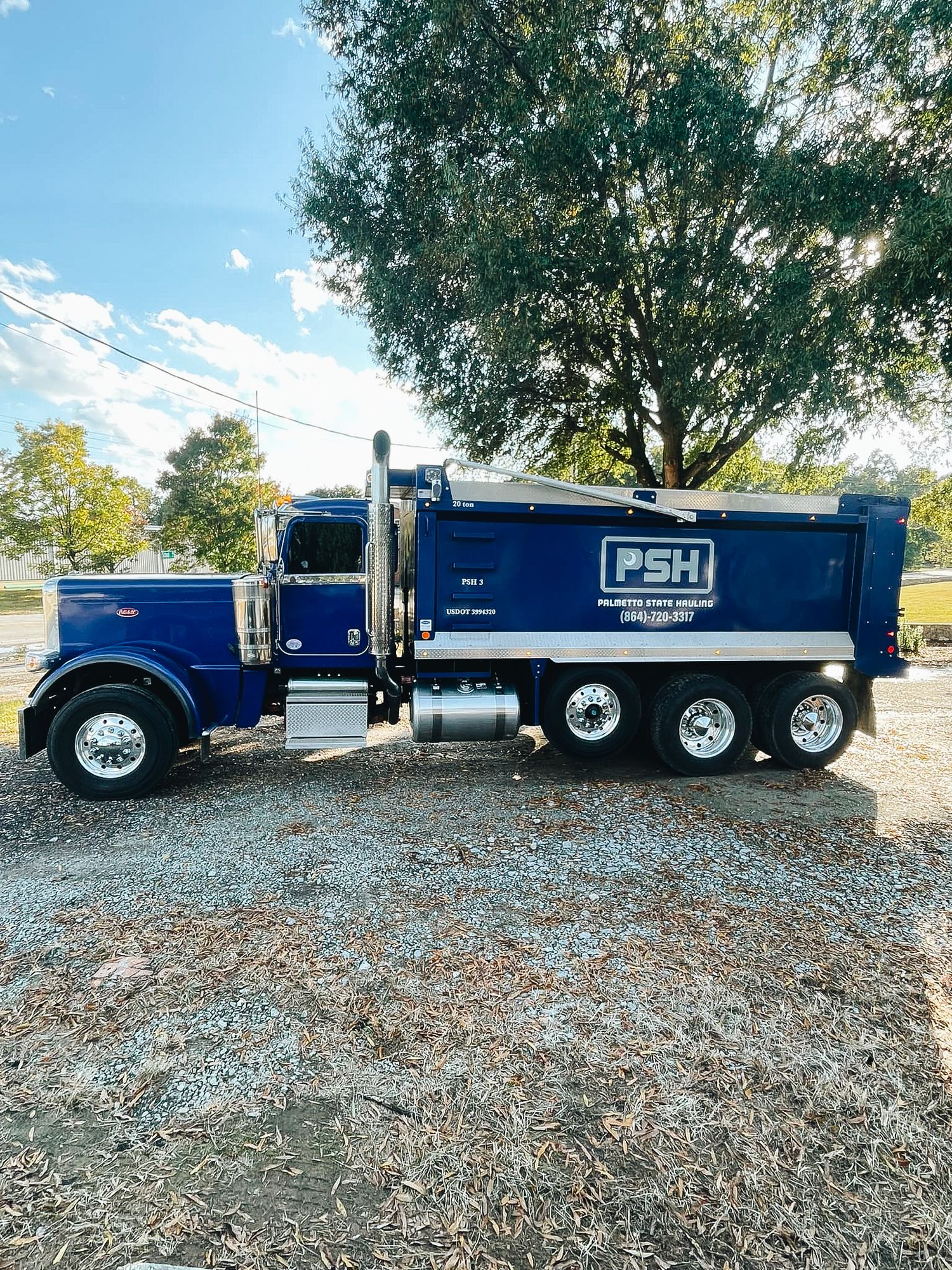 Blue PSH dump truck parked on gravel under trees against blue sky.