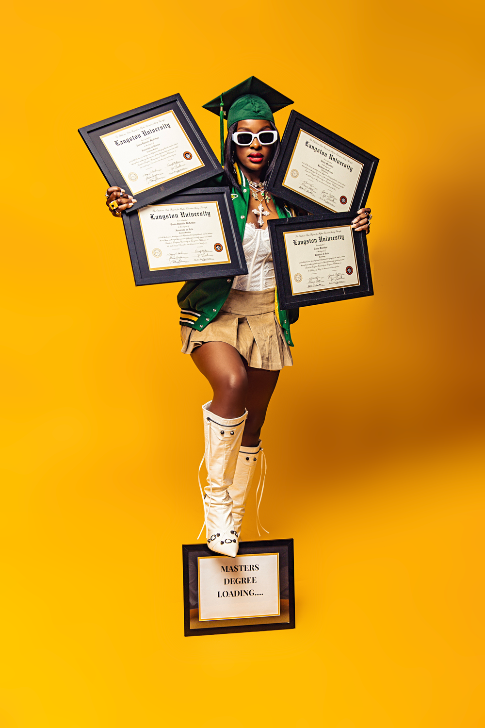 Graduate in green cap and gown shows off multiple diplomas against vibrant yellow background while wearing white boots.