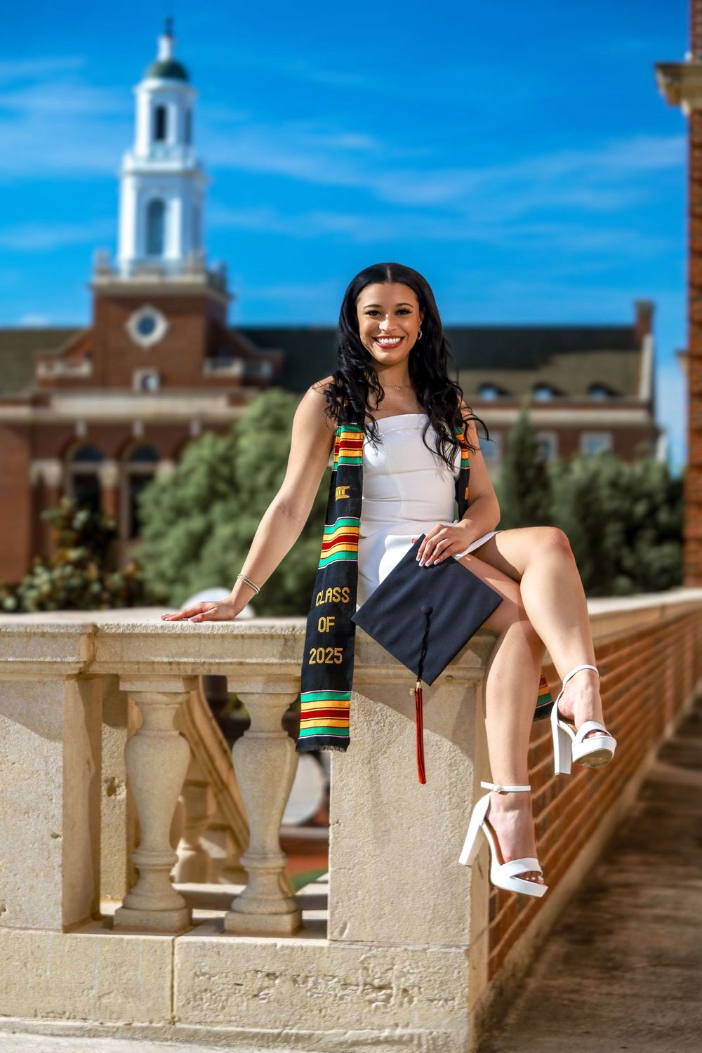 A graduate in a white dress and decorative sash poses on a stone balustrade with a stately brick building and bell tower behind.
