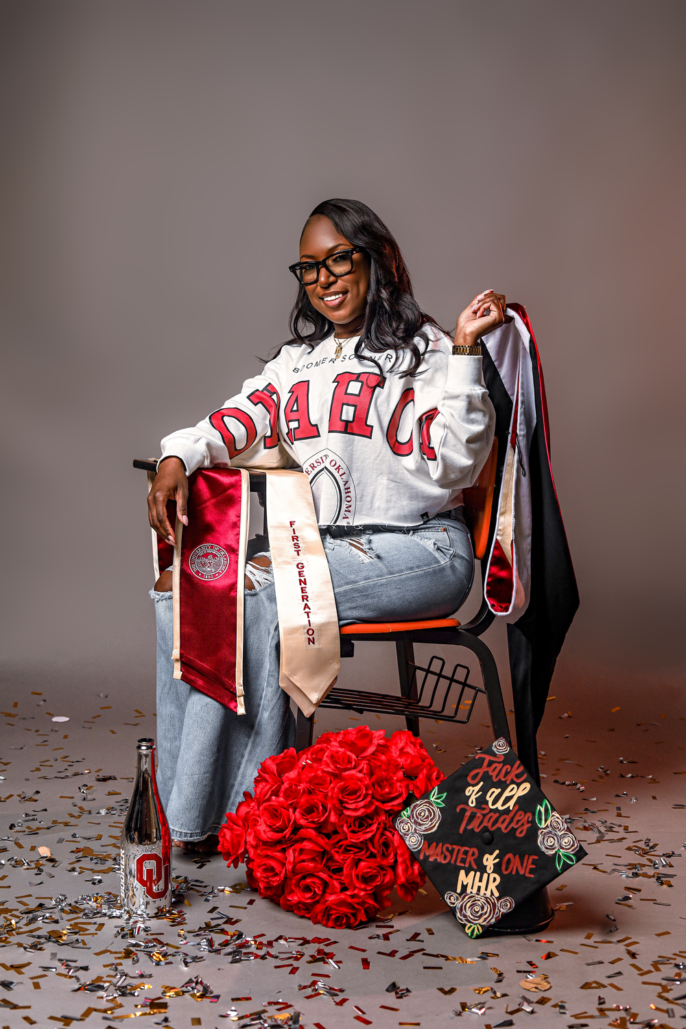 A person sits in a chair surrounded by red roses and graduation memorabilia while wearing a university letterman jacket.