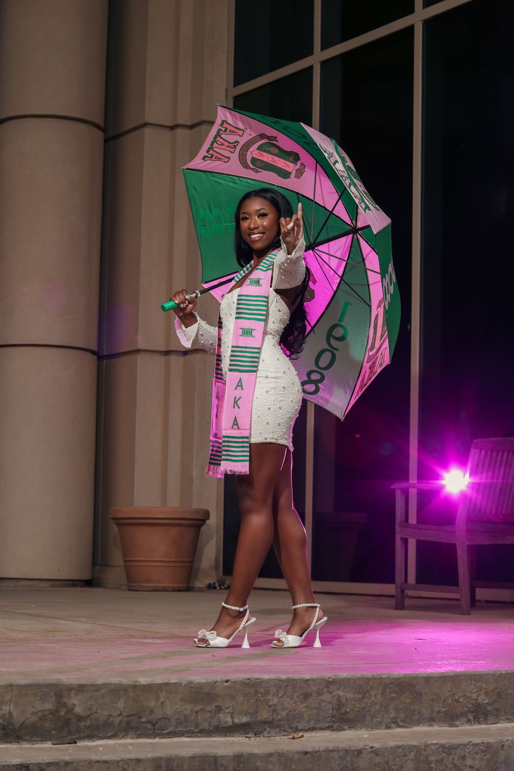A fashionable woman poses in a white dress with a pink and green umbrella at night under purple lighting.