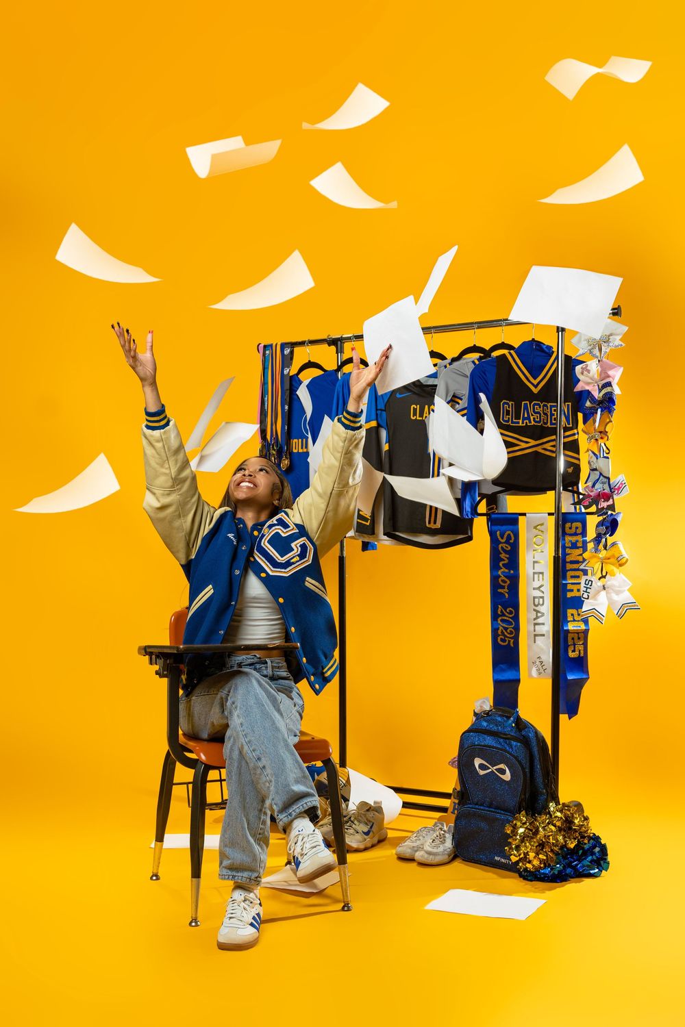 Student in blue varsity jacket celebrates by throwing papers in air against vibrant yellow background with backpack props.