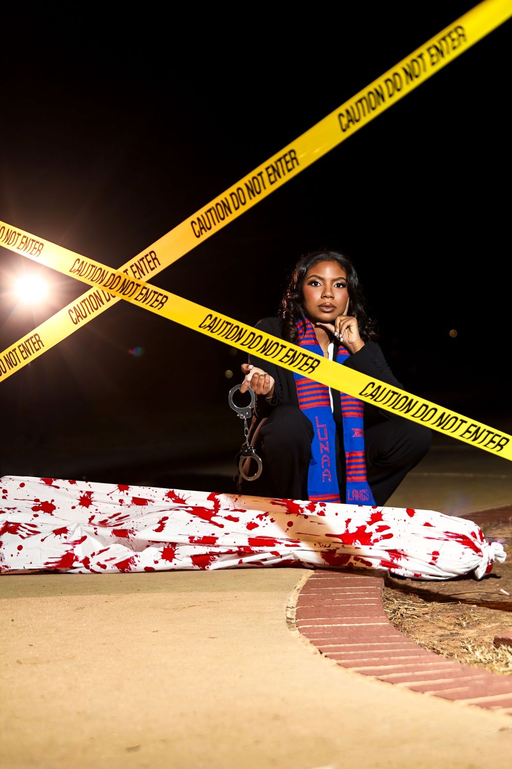 Crime scene photo with yellow caution tape and blood-spattered sheet behind police investigation barriers at night.