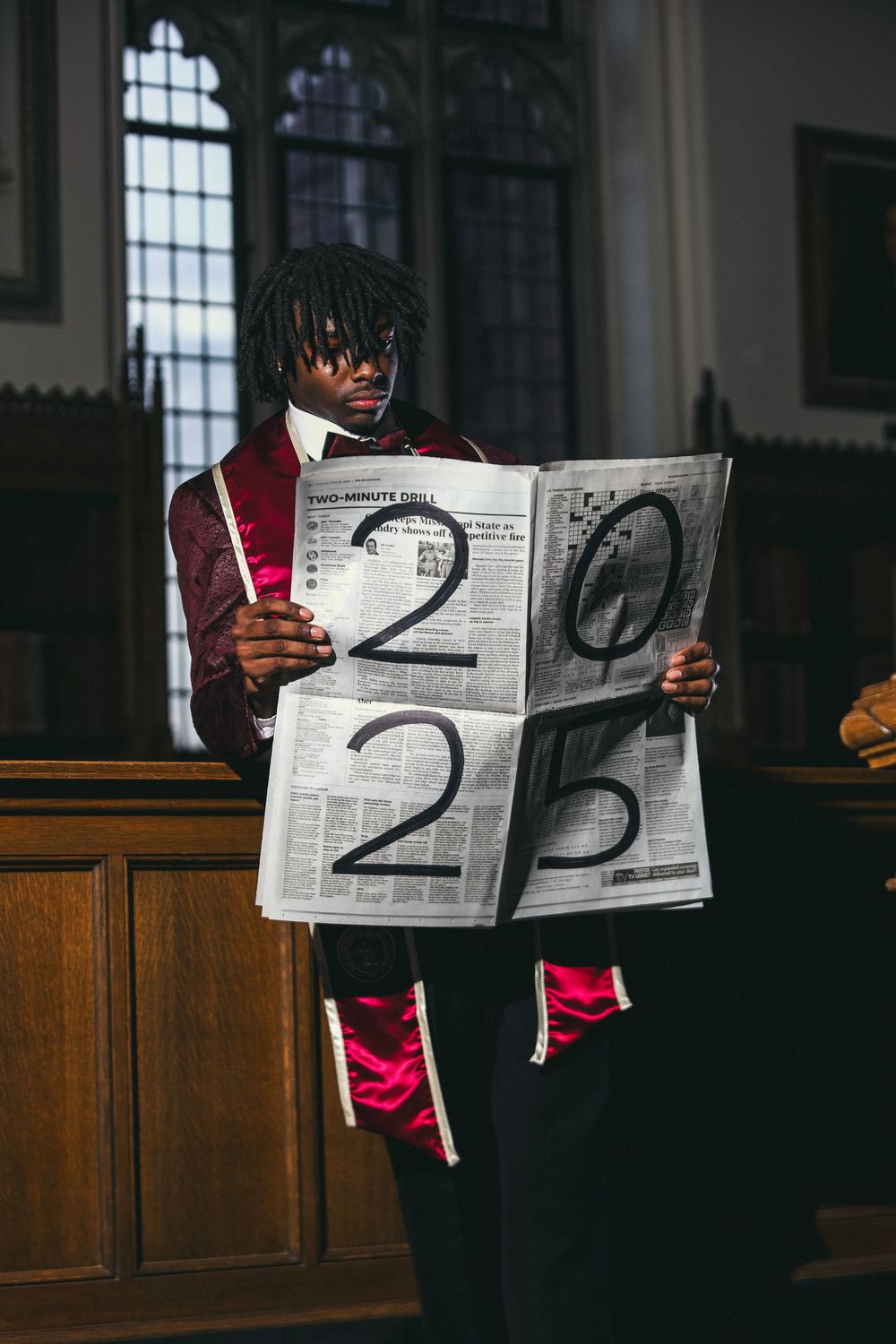 Student stands in church holding newspaper pages displaying "2025" written in thick black strokes.