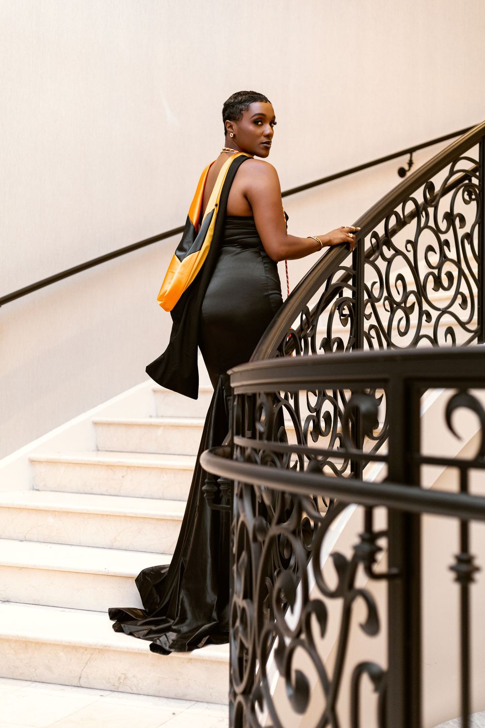A person in a black dress ascends an elegant staircase with ornate wrought iron railings while carrying a bag.