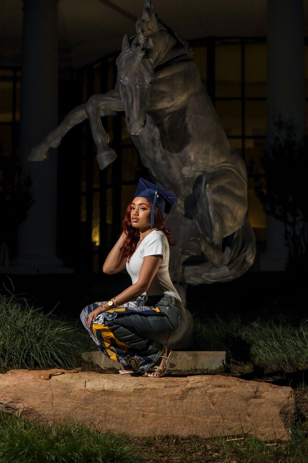 Portrait of a person in casual attire sitting near a large horse statue in an outdoor setting at night.