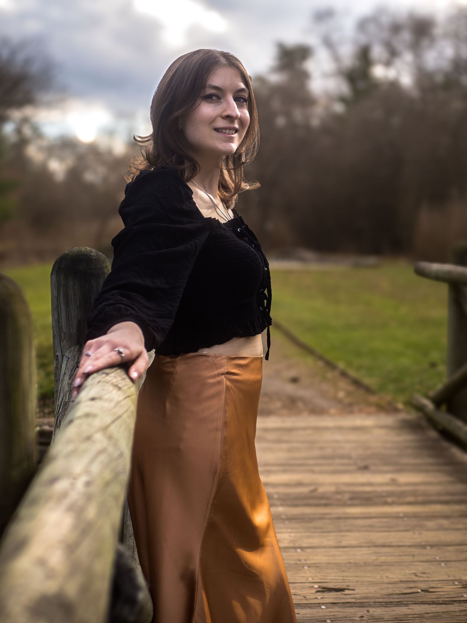 Woman in dark velvet top and silk skirt leans on rustic wooden bridge railing at dusk.