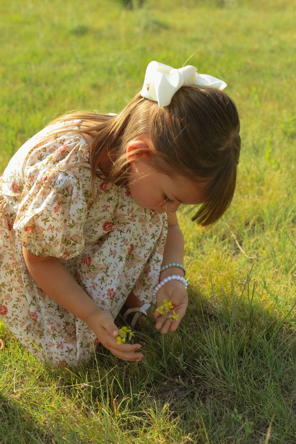 A person in a floral dress bends down to examine flowers in a sunlit grassy field.
