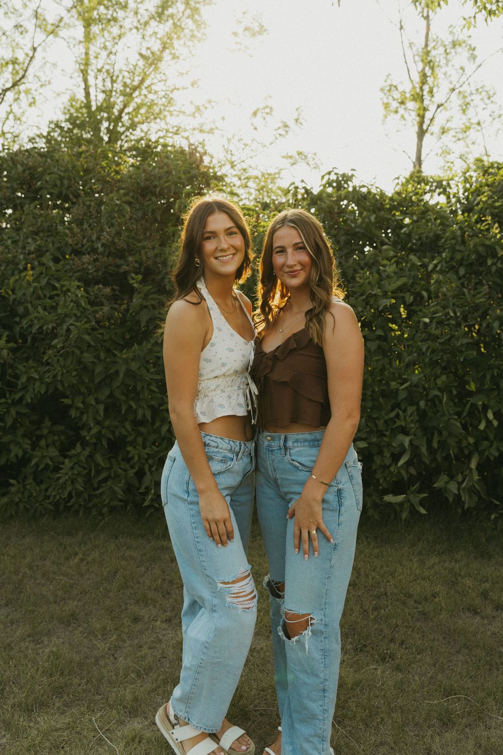 Two friends in ripped jeans and casual tops pose together against greenery at golden hour.