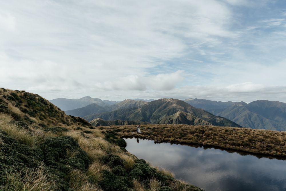 A serene mountain tarn reflects clouds and sky surrounded by tussock grass and rugged peaks in Wairarapa New Zealand by August and Vine