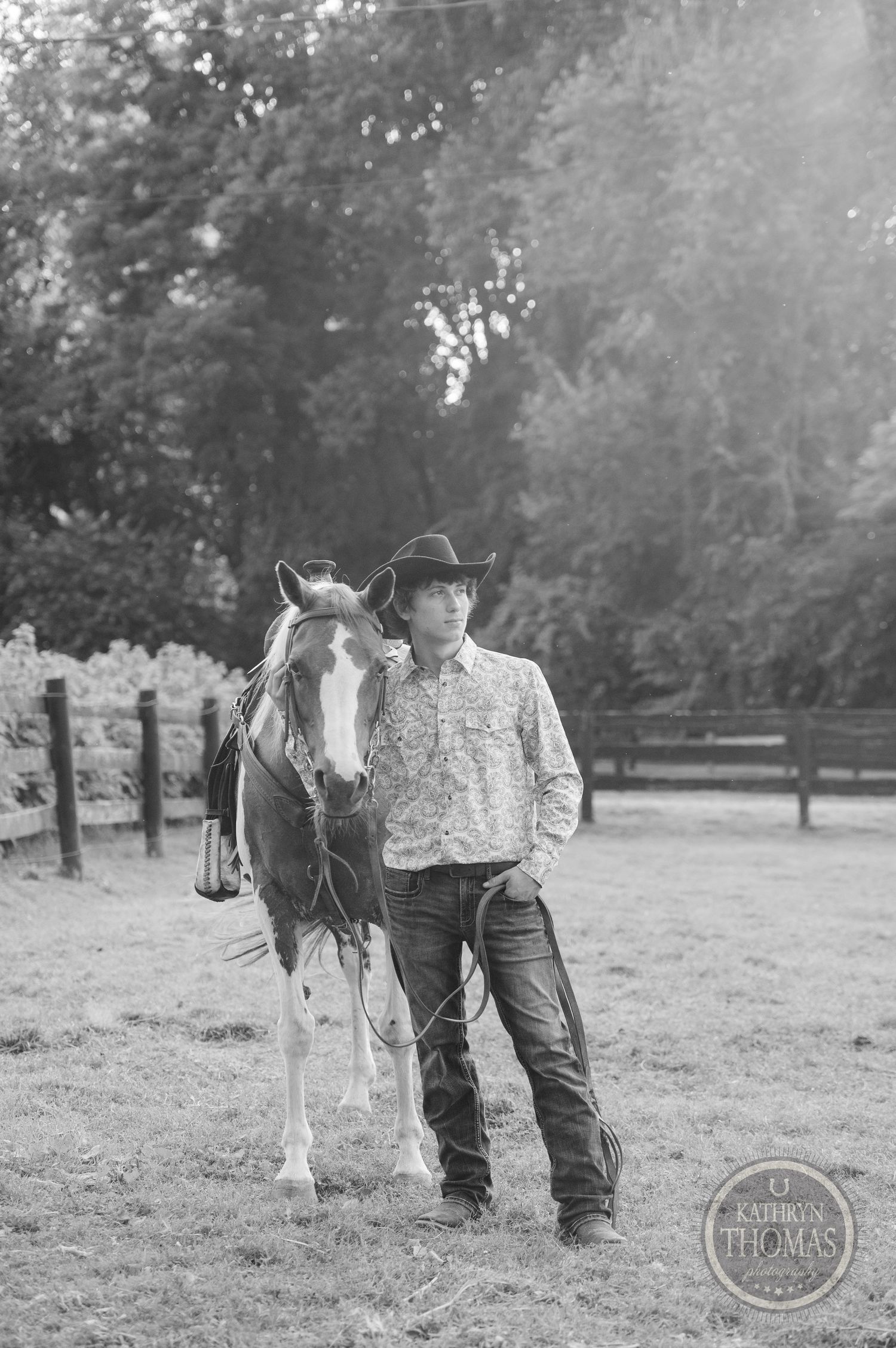 A black and white portrait of a western-dressed person with their paint horse in a paddock.