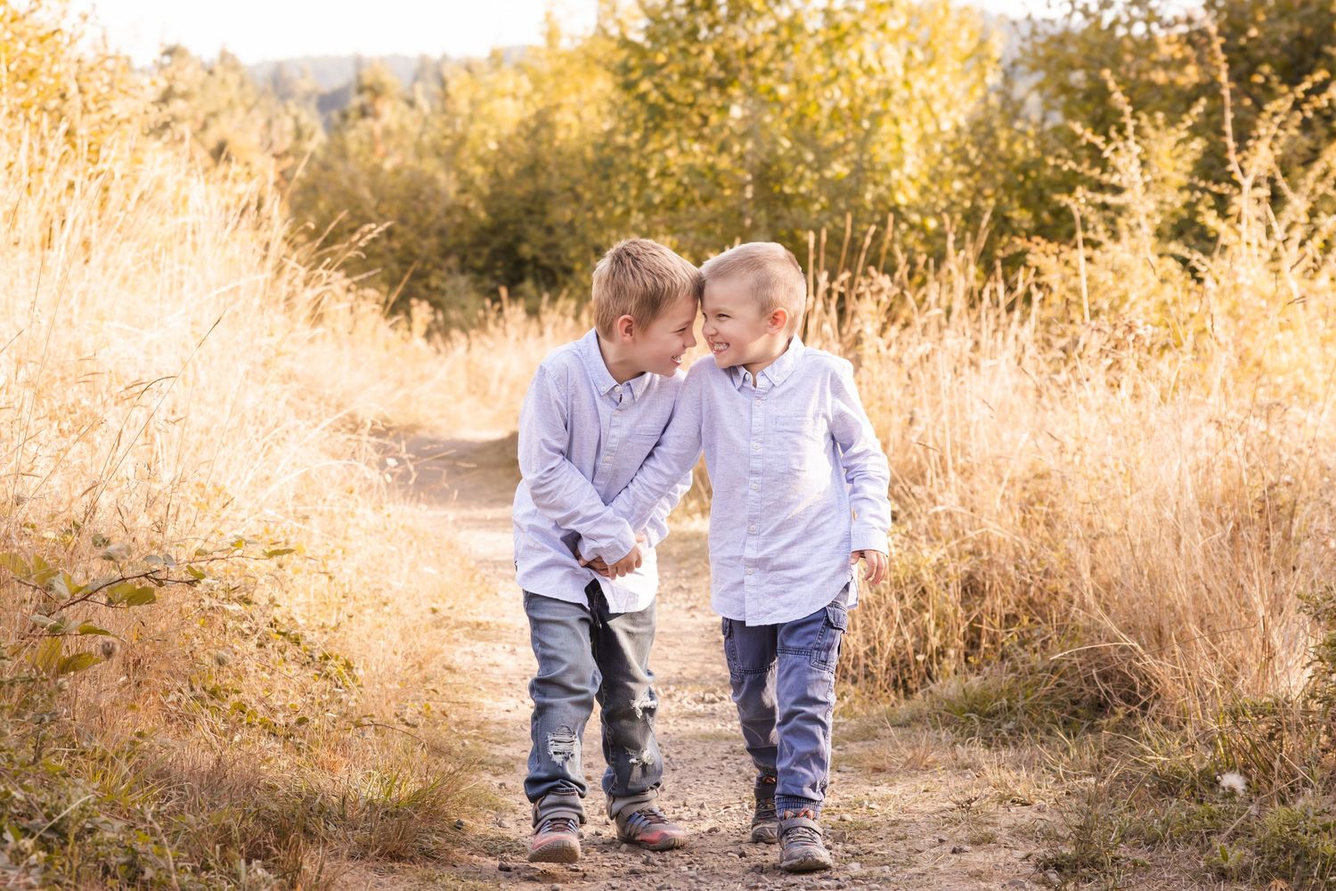 Two children in matching light shirts walk together on a dirt path surrounded by golden autumn grass at sunset.