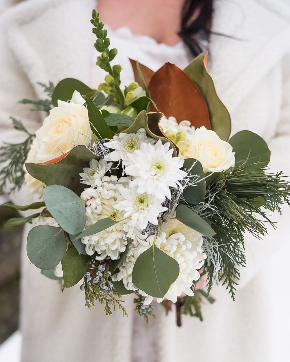 A bride, wearing a warm, fuzzy white jacket, holds her winter themed bouquet of flowers.