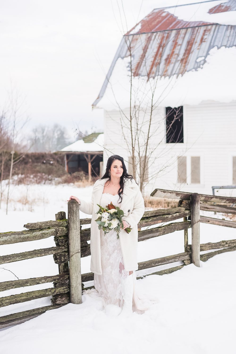 A bride stands near a fence in a snowy, winter wonderland, on her wedding day.