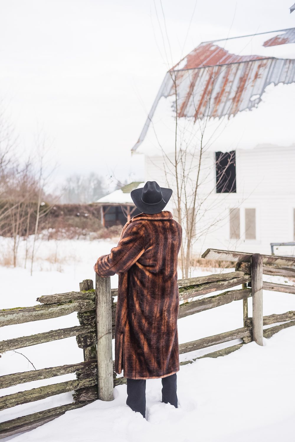 A groom stares off into the distance, past the snow covered barn, at his wedding venue.