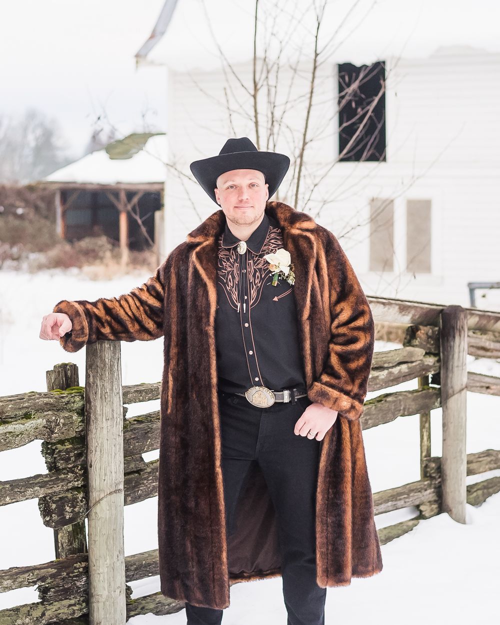 A groom poses outside in his faux fur jacket and cowboy hat.