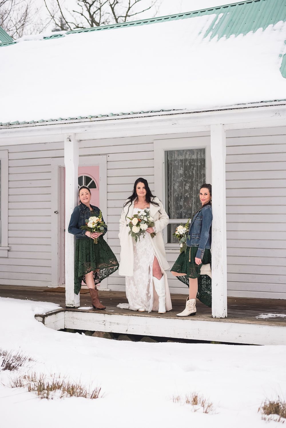 A bride, with her two bridesmaids, hold bouquets and pose for a portrait on the porch of a house in a snowy winter wonderland.